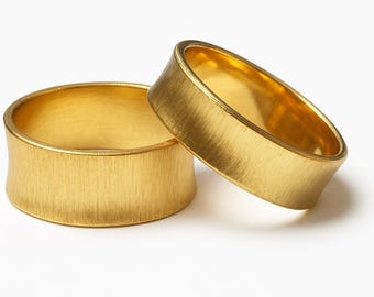 May include: Two wide gold bands with a brushed, matte finish on the exterior surface. The rings feature a slightly concave profile along the outer edge, showcasing a bright, polished interior surface that contrasts with the textured outer band.