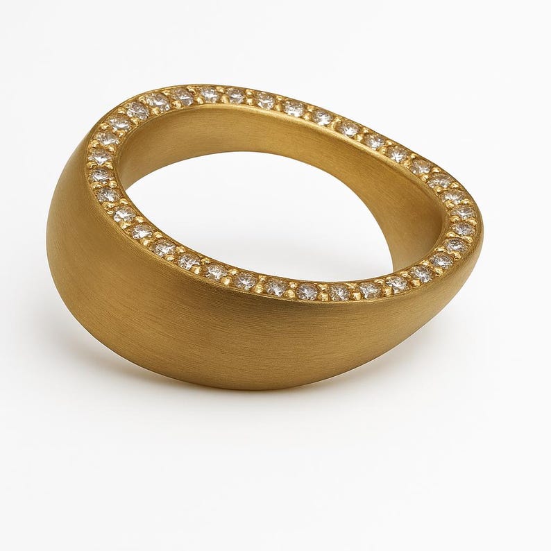 May include: A gold-colored ring with a smooth, matte finish. The ring is oval-shaped and features a row of small, sparkling gemstones set along the top edge. The ring is displayed against a white background.
