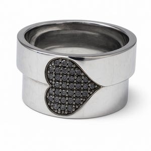 May include: A silver ring with a heart-shaped design filled with small, dark gemstones. The ring has a modern, minimalist style with a smooth, polished finish. The heart is centered on the ring's face, creating a striking visual contrast.
