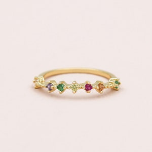 Pode incluir: Um anel dourado com uma série de pequenas pedras preciosas coloridas. As pedras preciosas estão dispostas em uma fileira, com cores como roxo, verde, vermelho e laranja. O anel está sobre um fundo rosa suave.