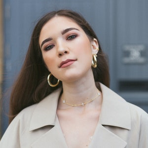 May include: A woman wearing gold hoop earrings and a gold butterfly necklace. She is also wearing a beige trench coat and a white lace top. Her hair is long and brown.