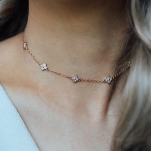 May include: Rose gold choker necklace featuring five clear, sparkling flower-shaped pendants. The delicate chain is visible against the skin, creating a subtle and elegant look. This necklace is a beautiful piece of jewelry.