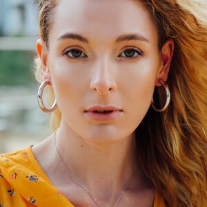 May include: A woman wearing silver hoop earrings and a delicate silver necklace with a small, heart-shaped pendant. She is wearing a yellow floral top. Her blonde hair is blowing in the wind.
