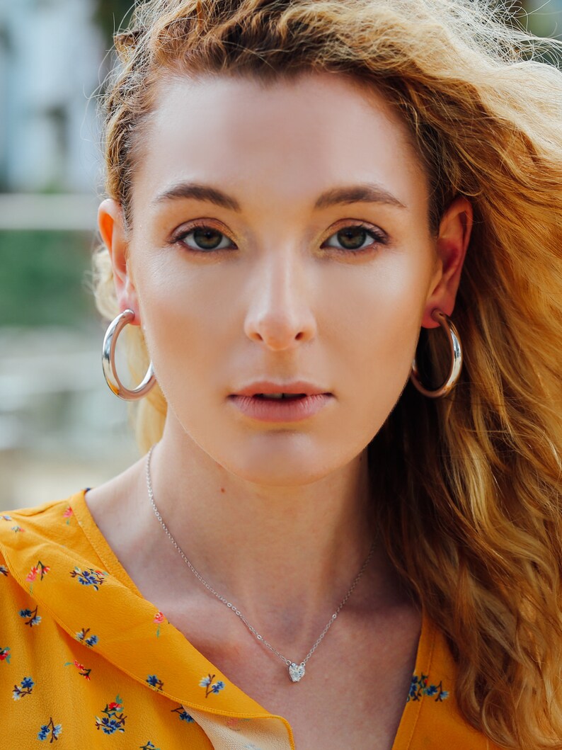 May include: A woman wearing silver hoop earrings and a delicate silver necklace with a small, heart-shaped pendant. She is wearing a yellow floral top. Her blonde hair is blowing in the wind.