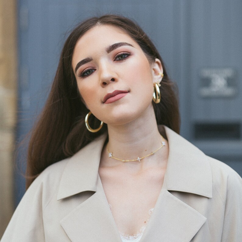 May include: A woman wearing gold hoop earrings and a gold butterfly necklace. She is also wearing a beige trench coat and a white lace top. Her hair is long and brown.