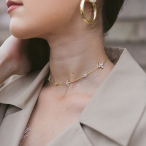 May include: A gold necklace with flower-shaped pendants and a pair of gold hoop earrings. The jewelry is worn with a beige trench coat. The earrings have a small, dangling charm with a blue center.
