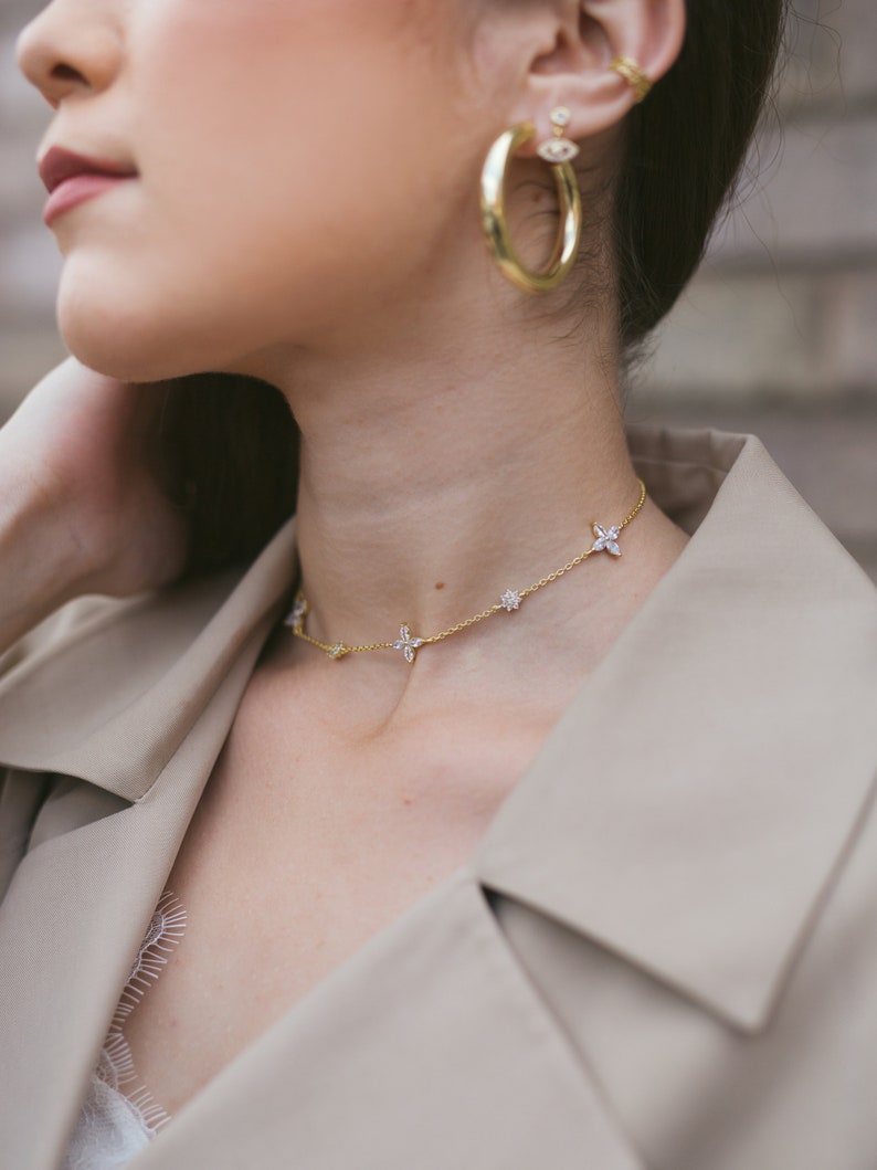 May include: A gold necklace with flower-shaped pendants and a pair of gold hoop earrings. The jewelry is worn with a beige trench coat. The earrings have a small, dangling charm with a blue center.
