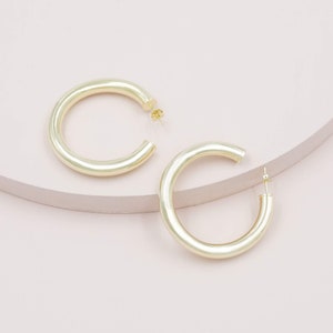 May include: Two gold-colored hoop earrings. The earrings are a thick, rounded design, with a post closure. The earrings are displayed on a light pink and beige surface. The earrings are a simple, yet elegant accessory.