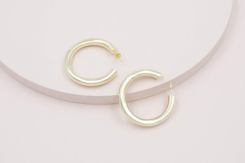May include: Two gold-colored hoop earrings. The earrings are a thick, rounded design, with a post closure. The earrings are displayed on a light pink and beige surface. The earrings are a simple, yet elegant accessory.