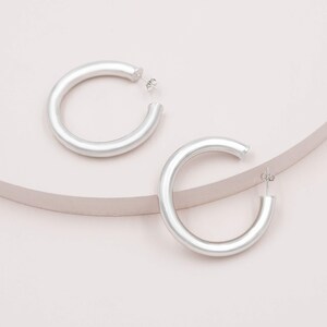 May include: Two silver-colored hoop earrings. The earrings are thick and have a smooth, matte finish. They are displayed on a light pink surface with a curved, light brown accent. The earrings are a simple, modern design.