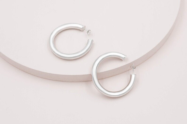 May include: Two silver-colored hoop earrings. The earrings are thick and have a smooth, matte finish. They are displayed on a light pink surface with a curved, light brown accent. The earrings are a simple, modern design.