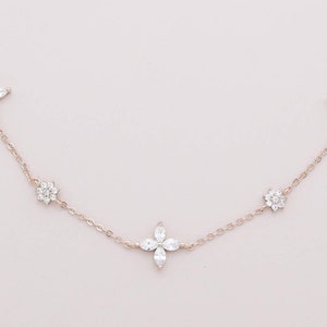 May include: A delicate necklace featuring a rose gold chain with five floral-shaped pendants. The pendants are adorned with clear, sparkling crystals, creating a refined and elegant accessory. The necklace is displayed against a soft pink background.
