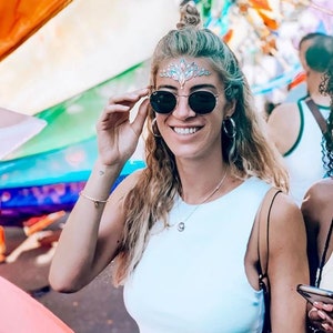 May include: A woman wearing round black sunglasses, a white tank top, and a decorative forehead jewel. She is smiling and holding her sunglasses. The background includes colorful flags and other people.