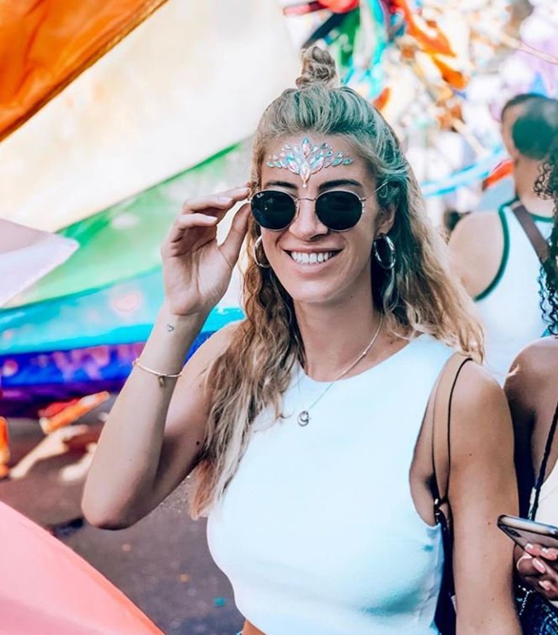 May include: A woman wearing round black sunglasses, a white tank top, and a decorative forehead jewel. She is smiling and holding her sunglasses. The background includes colorful flags and other people.