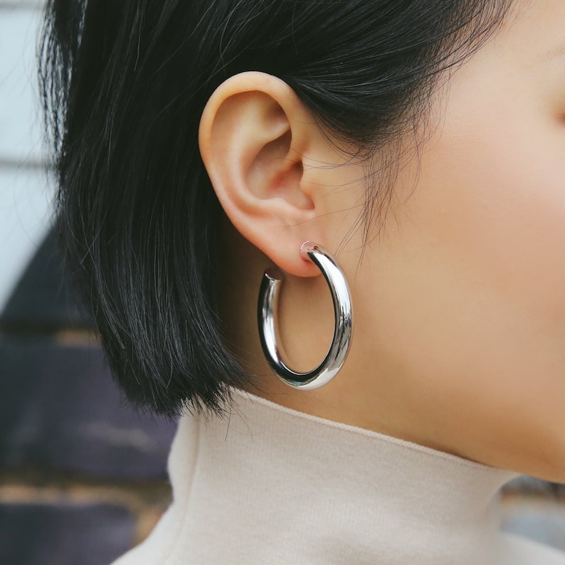 May include: A close-up of a person wearing a pair of large, shiny, silver hoop earrings. The earrings are thick and have a smooth, polished surface. The person has short, dark hair and is wearing a light-colored turtleneck.