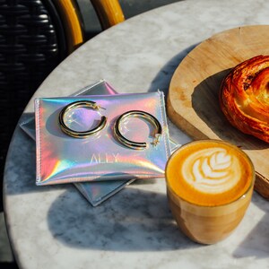 May include: A pair of gold hoop earrings rests on a holographic pouch with the word "ALLY" printed on it. A cup of latte with latte art and a pastry are also on the table. The earrings are thick and circular.