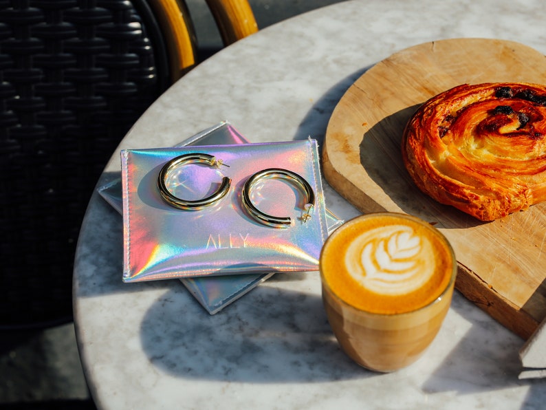 May include: A pair of gold hoop earrings rests on a holographic pouch with the word "ALLY" printed on it. A cup of latte with latte art and a pastry are also on the table. The earrings are thick and circular.