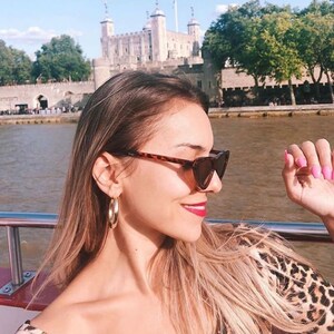 May include: A woman wearing tortoiseshell sunglasses and gold hoop earrings, with a leopard print top. The Tower of London is visible in the background, with the Thames River in front.