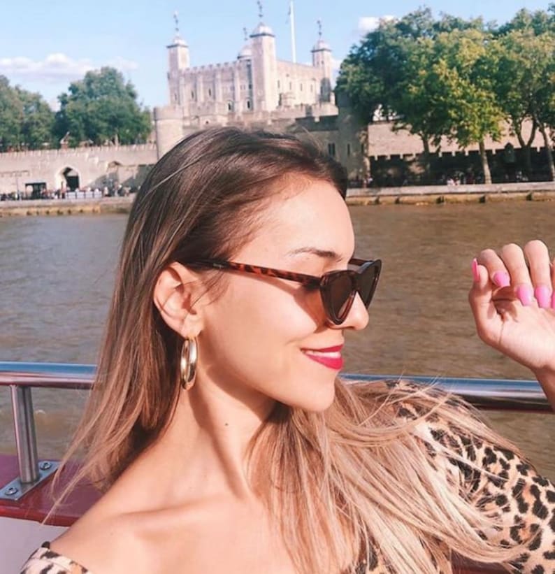 May include: A woman wearing tortoiseshell sunglasses and gold hoop earrings, with a leopard print top. The Tower of London is visible in the background, with the Thames River in front.