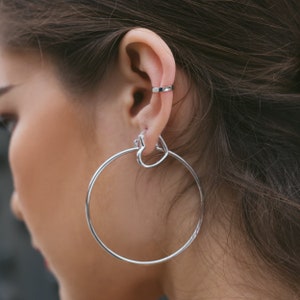 May include: Silver-toned hoop earrings with a heart-shaped accent and a matching ear cuff. The large, circular earrings are paired with a simple, geometric ear cuff, creating a modern jewelry set. The jewelry is shown on a person's ear.