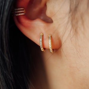 May include: Close-up of a person's ear adorned with gold earrings and a matching ear cuff. The earrings are small, hoop-style, and encrusted with clear gemstones. The ear cuff features three rows of similar gemstones.