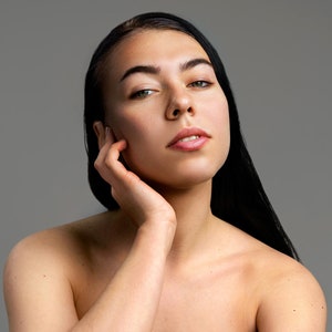 Peut inclure: Une femme aux longs cheveux noirs, sans v&ecirc;tements, regarde la cam&eacute;ra avec une expression douce. Sa main repose sur sa joue.