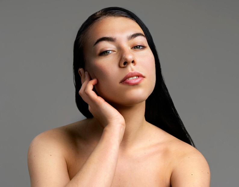 Peut inclure: Une femme aux longs cheveux noirs, sans v&ecirc;tements, regarde la cam&eacute;ra avec une expression douce. Sa main repose sur sa joue.