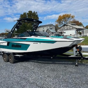 May include: A white and teal Malibu wakeboard boat on a black trailer with black wheels. The boat has a black tower with a black canvas top. The boat is parked on a gravel surface.
