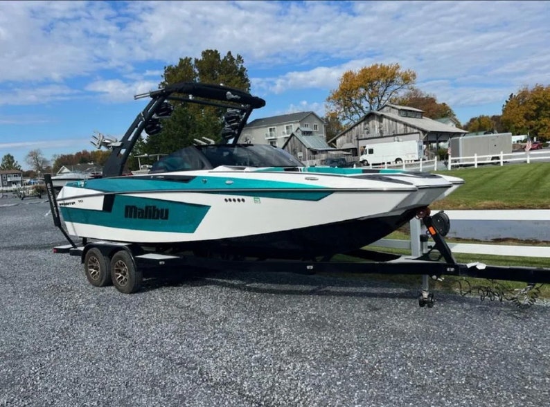 May include: A white and teal Malibu wakeboard boat on a black trailer with black wheels. The boat has a black tower with a black canvas top. The boat is parked on a gravel surface.