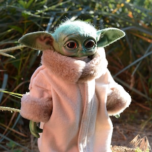 May include: A small green plush toy with large ears and big eyes wearing a white faux fur coat. The toy is sitting on a tree stump.