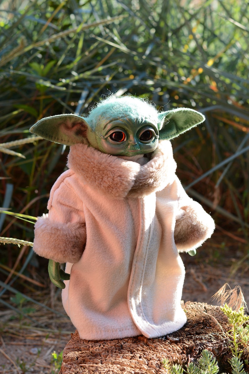 May include: A small green plush toy with large ears and big eyes wearing a white faux fur coat. The toy is sitting on a tree stump.
