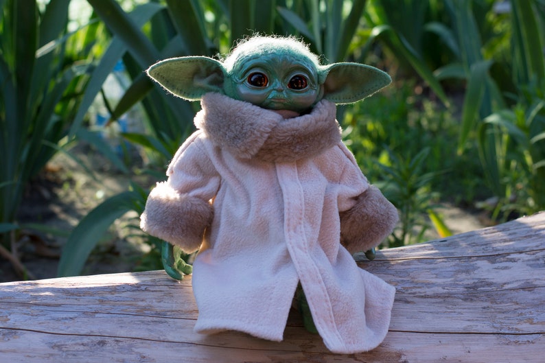 May include: A green plush toy with large ears and brown eyes wearing a white coat with a brown fur collar and cuffs. The toy is sitting on a wooden log.
