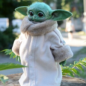 May include: A green plush toy with large ears wearing a white coat with a brown fur collar and cuffs. The toy is standing on a tree stump.