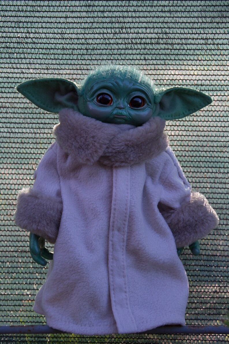 May include: A plush toy of a green alien character wearing a white coat with a fur collar and cuffs. The character has large ears and big brown eyes.