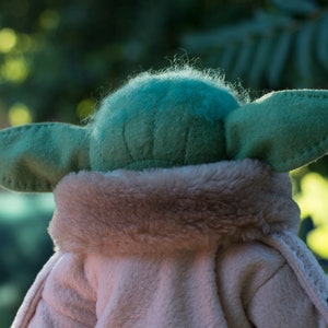 May include: A plush toy of a green character with large ears, wearing a brown faux fur coat. The toy is facing away from the camera.