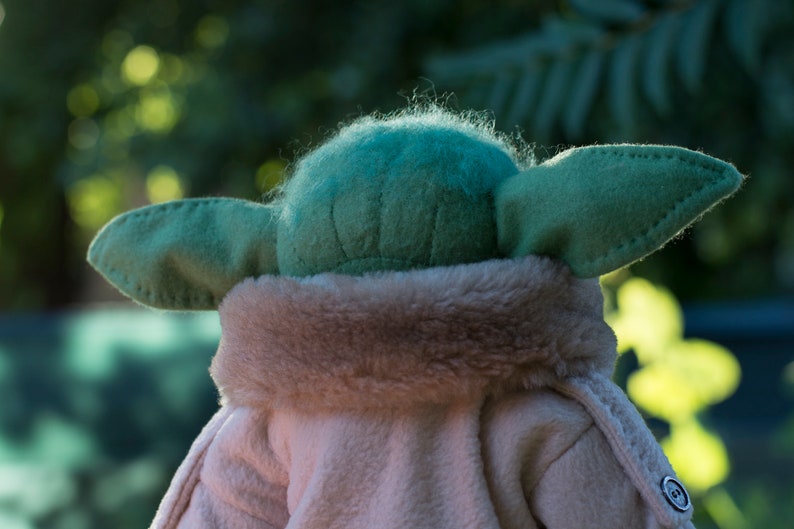 May include: A plush toy of a green character with large ears, wearing a brown faux fur coat. The toy is facing away from the camera.