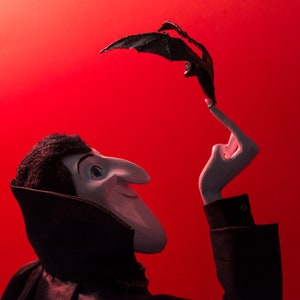 May include: A black and white puppet of a vampire with a black cape and a bat puppet in its hand against a red background.