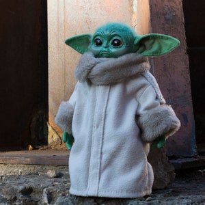 May include: A green and brown plush toy wearing a white coat with a fur trim. The toy has large ears and big eyes.