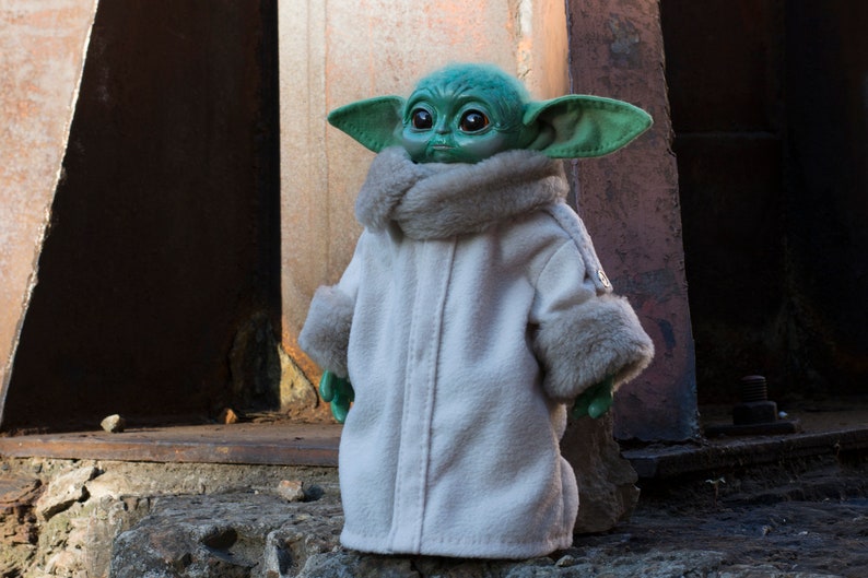 May include: A green and brown plush toy wearing a white coat with a fur trim. The toy has large ears and big eyes.