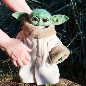 May include: A green plush toy with large eyes and a white coat, standing on a tree stump. The toy is wearing a white coat with a brown fur collar and cuffs.