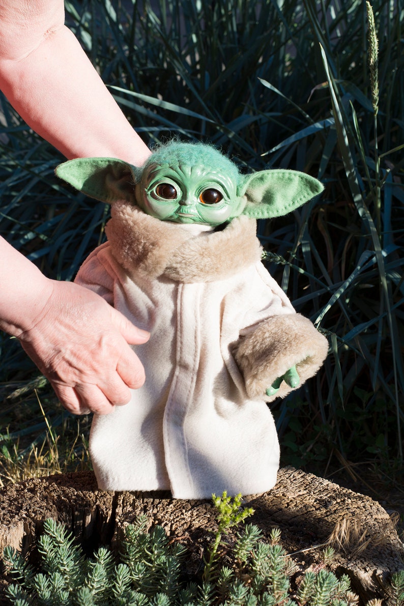 May include: A green plush toy with large eyes and a white coat, standing on a tree stump. The toy is wearing a white coat with a brown fur collar and cuffs.