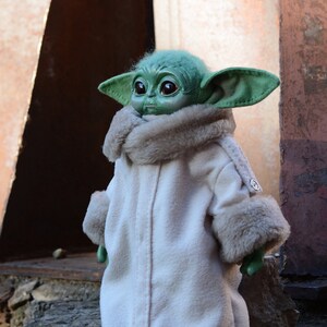 May include: A small green plush toy with large eyes and a white fur coat. The toy is standing on a rock.