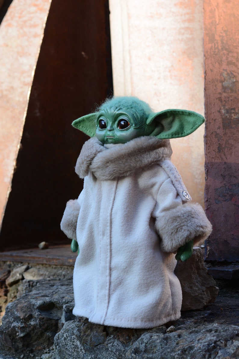 May include: A small green plush toy with large eyes and a white fur coat. The toy is standing on a rock.