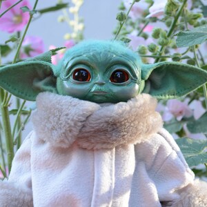 May include: A green plush toy with large eyes and a furry white collar. The toy is posed in a garden setting with pink flowers.