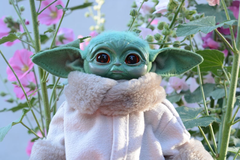 May include: A green plush toy with large eyes and a furry white collar. The toy is posed in a garden setting with pink flowers.