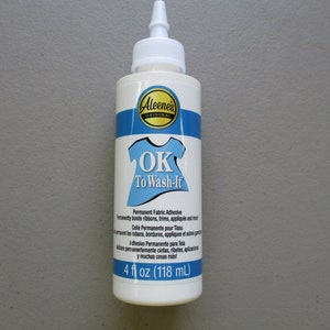 May include: A white bottle of Aleene's Original OK To Wash-It permanent fabric adhesive. The bottle has a blue and white label with the text "OK To Wash-It" and a graphic of a t-shirt. The bottle contains 4 fl oz (118 mL).
