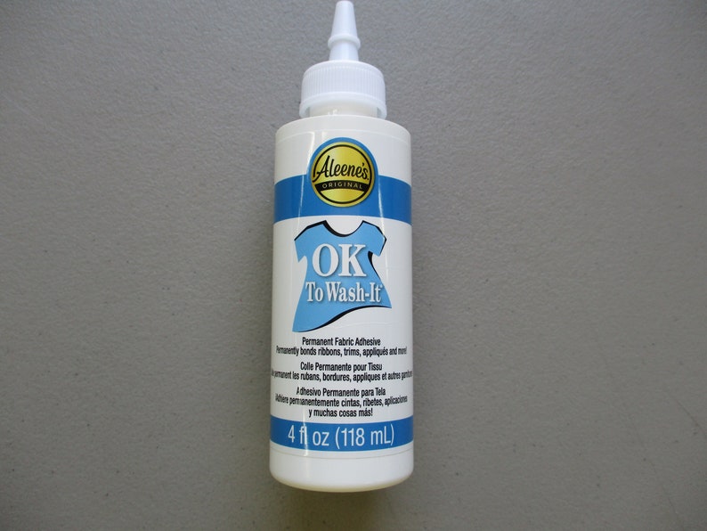 May include: A white bottle of Aleene's Original OK To Wash-It permanent fabric adhesive. The bottle has a blue and white label with the text "OK To Wash-It" and a graphic of a t-shirt. The bottle contains 4 fl oz (118 mL).