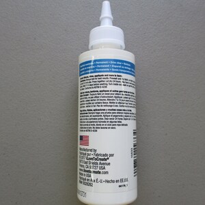 May include: A white bottle of fabric adhesive with a pointed applicator tip. The label includes text in English and Spanish, with the American flag visible. The bottle is designed for fabric applications.