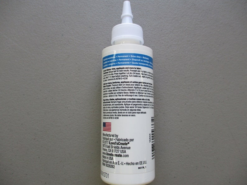 May include: A white bottle of fabric adhesive with a pointed applicator tip. The label includes text in English and Spanish, with the American flag visible. The bottle is designed for fabric applications.