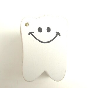 May include: A white, tooth-shaped wooden ornament with a smiling face. The tooth has a black outline and features two black dots for eyes and a curved line for a mouth. A small gold-colored metal eyelet is at the top.